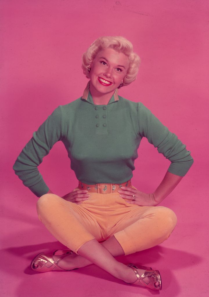 Full-length studio portrait of American actor and singer Doris Day sitting on the floor in front of a pink backdrop, her legs crossed and her hands on her hips. Day is smiling and wearing a long sleeve blue shirt, yellow capri pants, and gold shoes.