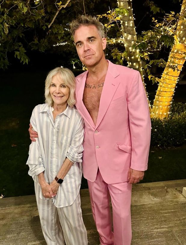 An elderly woman standing next to Robbie Williams, who wears a pink suit