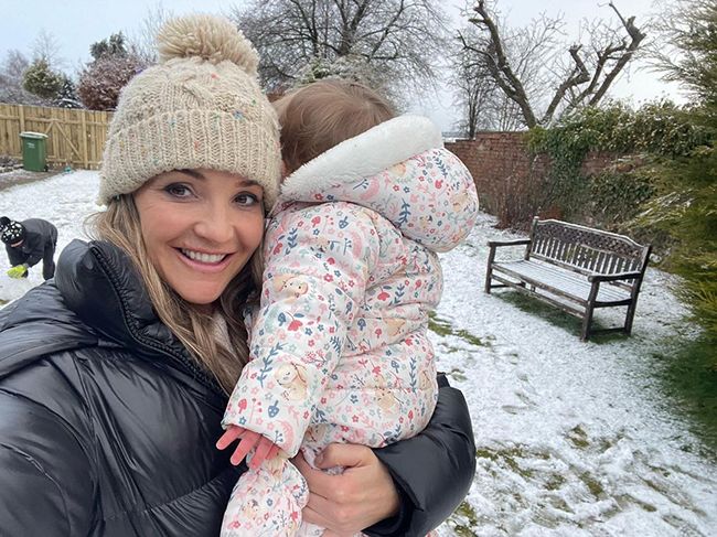 helen skelton in padded jacket and wooly hat standing in snowy garden holding baby daughter elsie