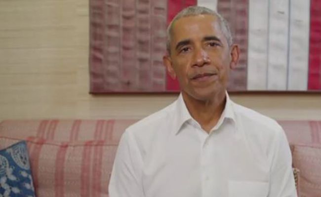barack obama wearing a white shirt on a cream and salon hued striped sofa with contrasting blue patterned cushion and a complementary pink hued wall hanging in the background