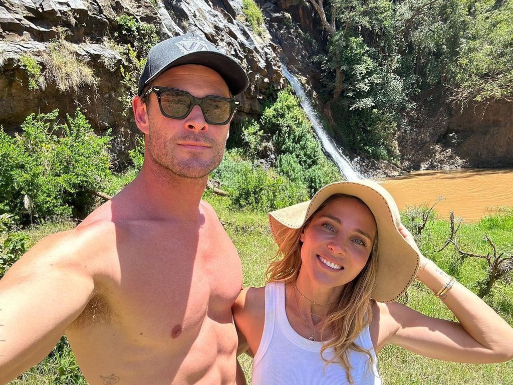 Chris and Elsa smiling for a selfie near a waterfall, both are wearing hats and her is wearing sunglasses