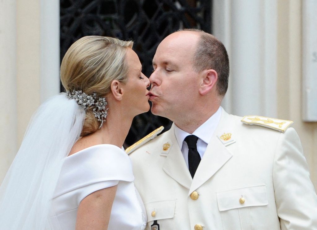 Princesa Charlene em um vestido de noiva beijando o príncipe Albert que usa um terno branco