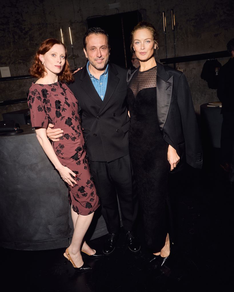 Karen Elson, Piergiorgio del Moro et Carolyn Murphy sourient devant la caméra. Karen porte une robe rouge avec des détails floraux noirs et Carolyn porte une robe noire avec un blazer surdimensionné.