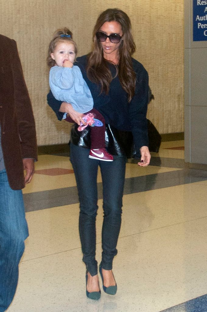 Harper Beckham wore a blue top, maroon leggings, and Nike sneakers. She was carried by Victoria Beckham, who dressed in black skinnies and heels. Both looked fashionable while traveling.