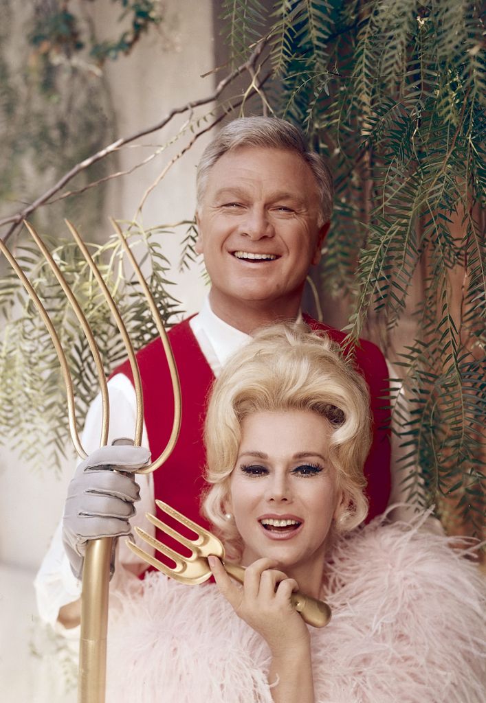 American actor Eddie Albert (1906 - 2005) holds a gold colored pitchfork and stands behind his tv wife Hungarian-born actress Eva Gabor (1919 - 1995), who holds a gold colored hand rake, as they pose for a publicity portrait 