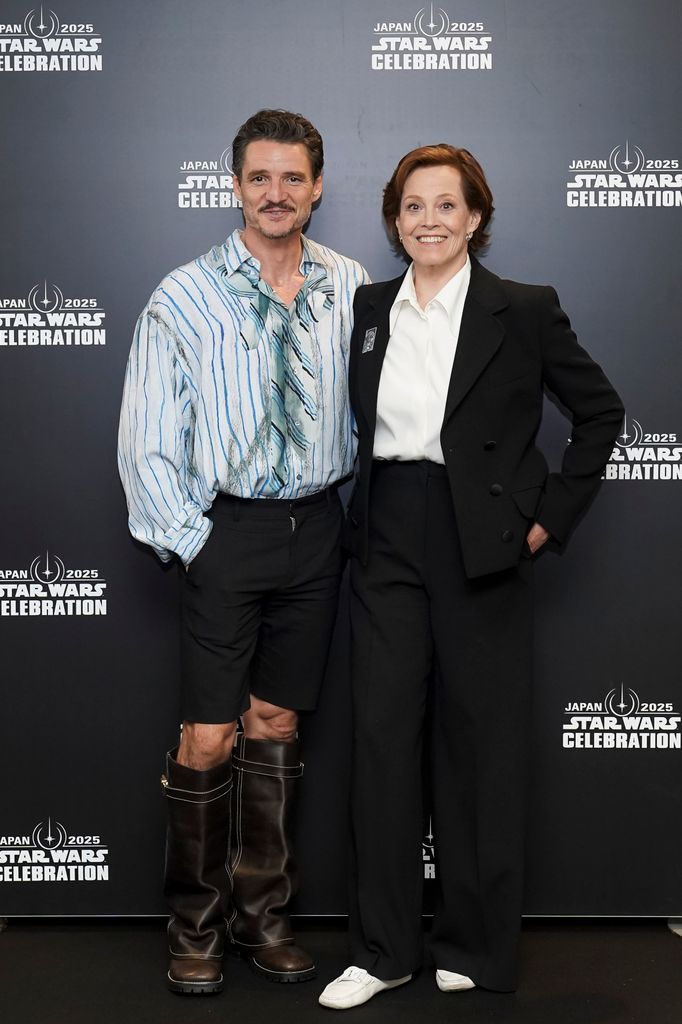 Pascal stands smiling with Sigourney Weaver in front of a matte black backdrop emblazoned with “Japan 2025 Star Wars Celebration” logos. He’s wearing tailored black shorts, a voluminous button-down shirt with painterly blue-and-aqua vertical stripes, and tall, heavy-duty brown leather boots that climb nearly to his knees. The boots are the centerpiece—thick-soled, overbuilt, and exaggerated in proportion. 