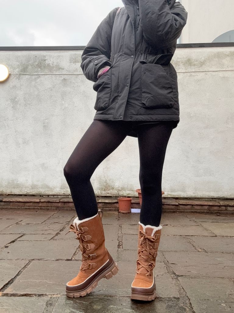 A cropped image of a woman in a black puffer coat, black shorts and tights with knee high hiking boots with thick soles and shearling lining