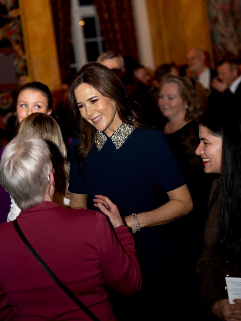 Queen Mary in dress with glitter collar talking to people in dark room