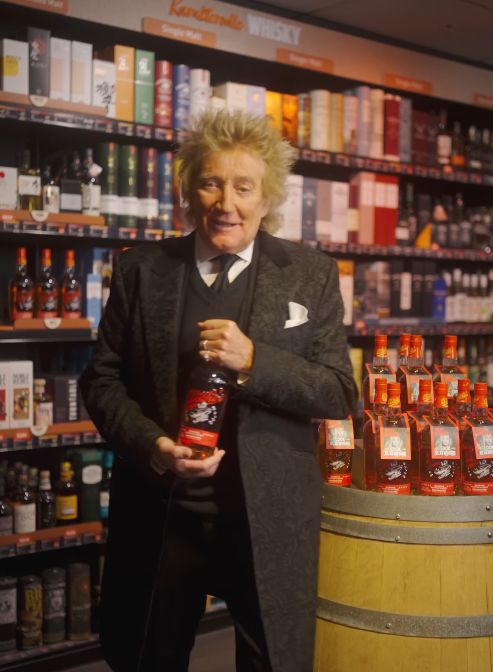 Rod Stewart in a shop holding a bottle of whisky