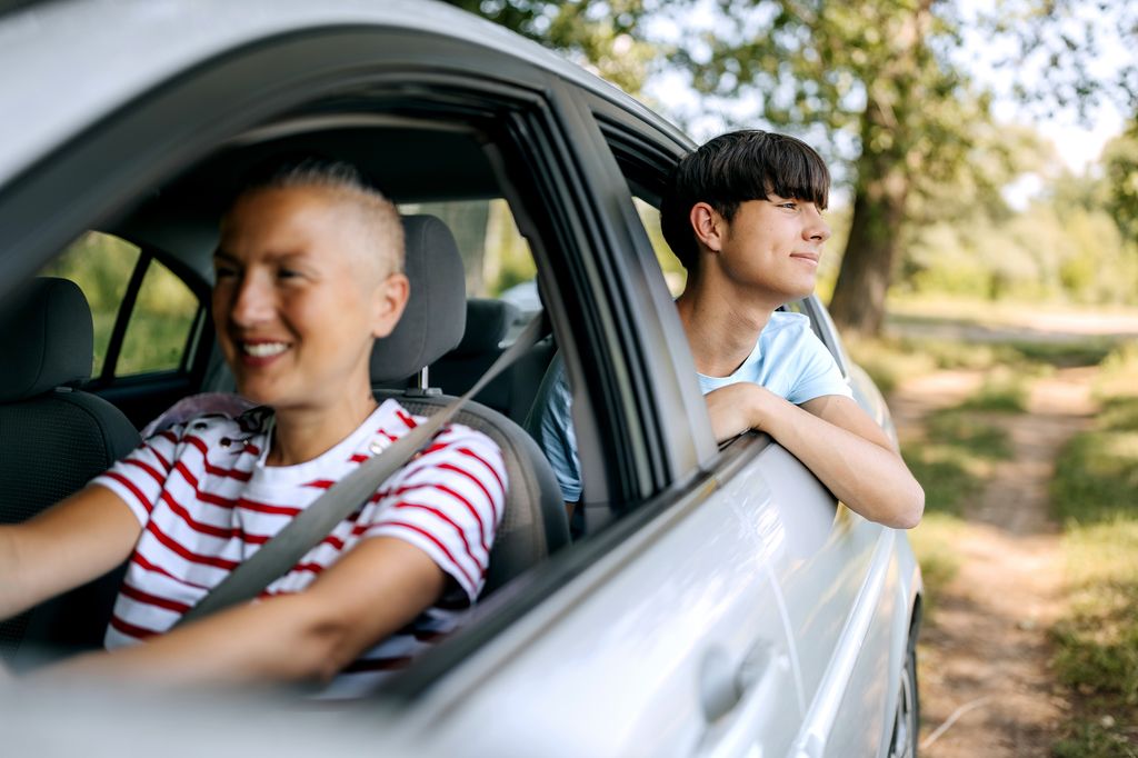 A teenage boy leaning through the open back window of a moving car, smiling and enjoying the fresh air and surrounding nature
