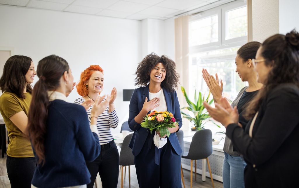 Smiling female professional receiving flowers from coworkers at office. Multi-ethnic business women celebrating an achievement of a colleague at start up office.