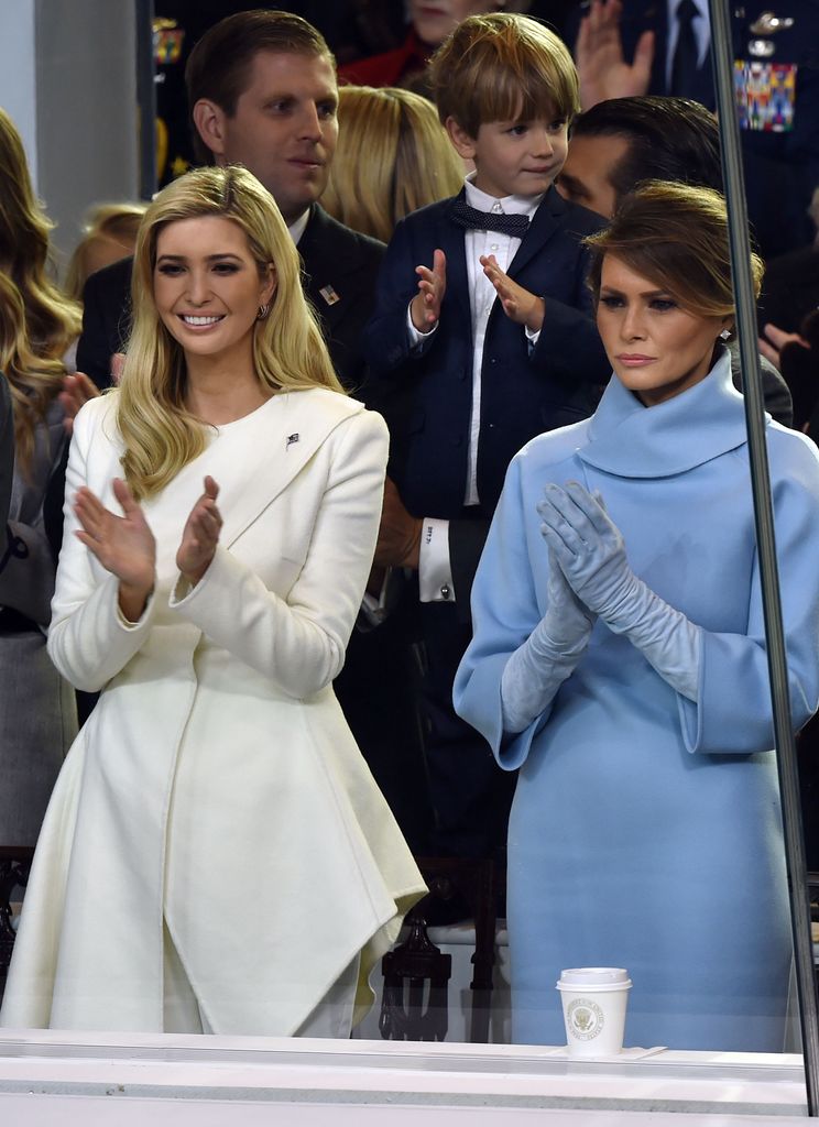 Wearing a structured white coat with asymmetrical lines, Ivanka smiles as she claps. Next to her Melania in light blue. Both exude elegance in contrasting pastel tones at the Presidential Inauguration Parade on January 20, 2017 in Washington