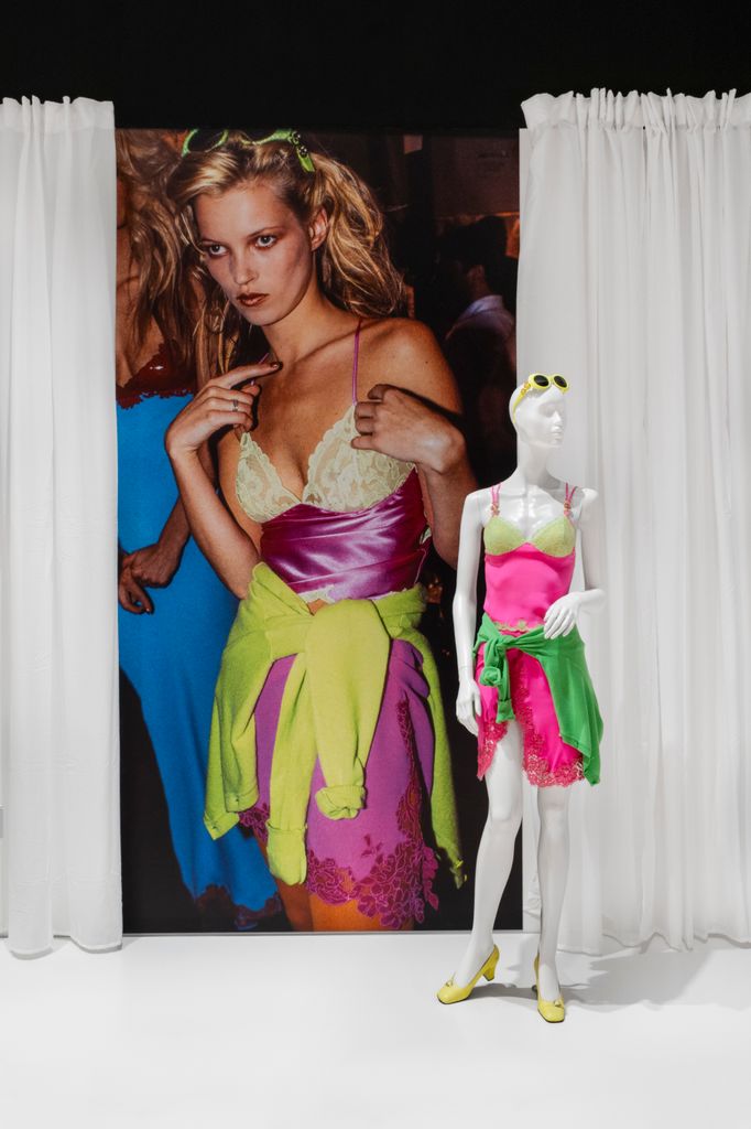 A mannequin wears a bright pink and lime green lace-trimmed Versace slip dress with a green sweater tied at the waist, styled exactly as worn by Kate Moss in the archival image enlarged behind, capturing the essence of '90s party style.