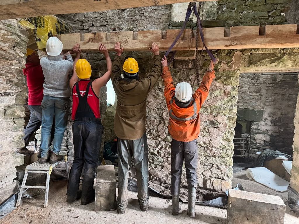 Five people holding up wooden beam inside farmhouse as construction team work on renovation