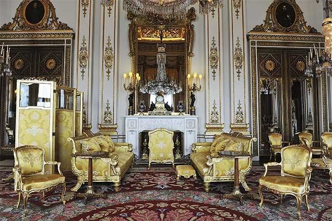a white walled room with exceptionally high ceilings is filled with gold chairs and sofas positioned throughout the room