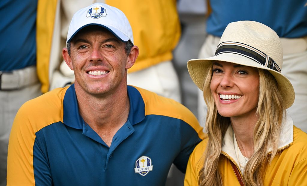Rory McIIroy smiling, wearing a white hat sitting next to his wife Erica, who is wearing a hat and smiling