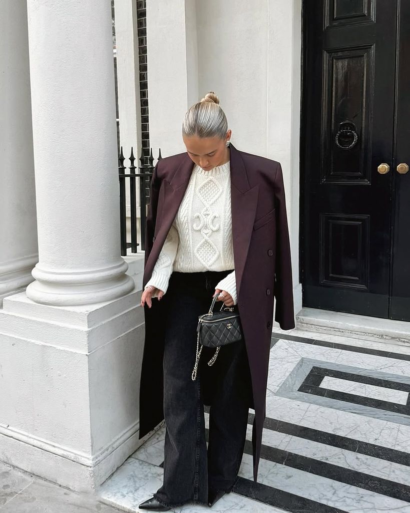 Molly-Mae stands in front of a black door and white columns, wearing a burgundy long coat draped over a chunky cream knit sweater, wide-leg black jeans, pointed boots, and carrying a quilted black Chanel mini bag. Her hair is slicked back into a sleek bun, completing the sophisticated look.