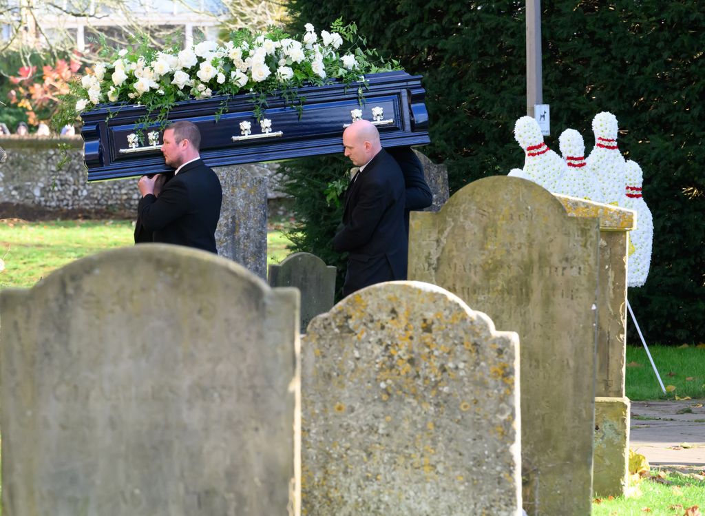 A coffin being carried past gravestones and a bowling pin floral tribute