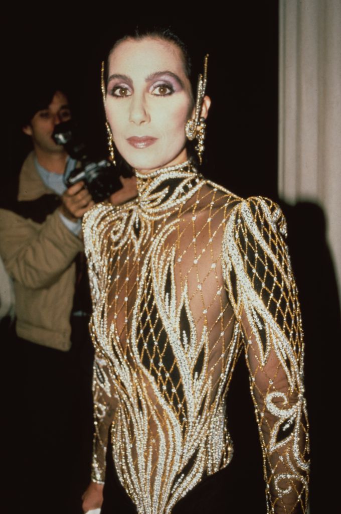 Cher stuns in a sheer beaded Bob Mackie gown at the Met Gala, wearing dramatic diamond chandelier earrings and matching hairline jewels in a celestial, body-hugging ensemble.