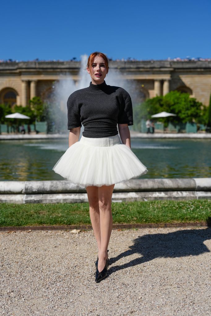  Emma Roberts wears a charcoal grey short-sleeved fitted knit top with a mock neck, tucked into a voluminous ivory tulle mini skirt