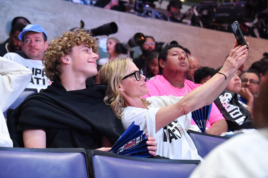 Julie Bowen tient un téléphone pour prendre un selfie alors qu'elle est assise parmi une foule de gens à côté de son fils Oliver qui sourit à la caméra.