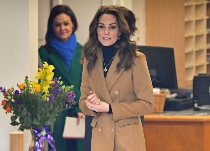 Kate Middleton in brown coat standing next to a vase of flowers