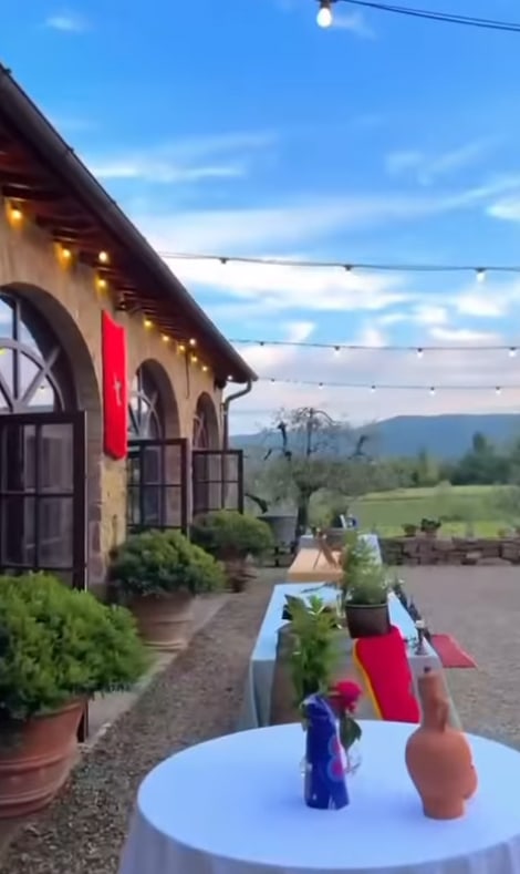 Outdoor event setup beside a rustic stone building with arched windows, flag hanging, string lights above, and tables decorated with bottles and clay vases. Green landscape and mountains visible in the background.