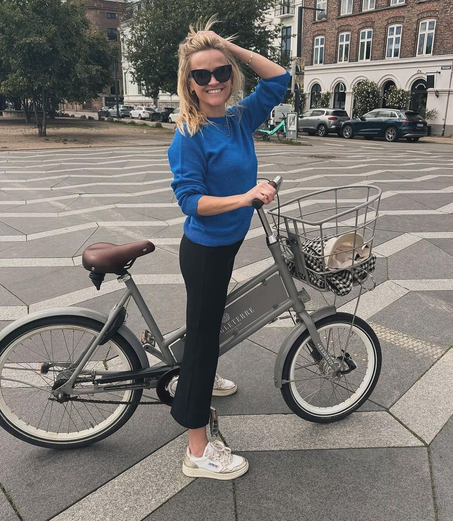 Reese Witherspoon straddles a bike in a town square