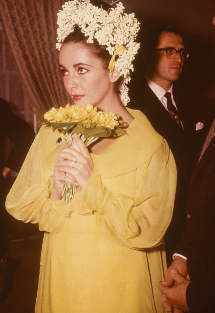 British-born actor Elizabeth Taylor, a yellow dress and floral headdress, holds a bouquet of flowers at her wedding to actor Richard Burton