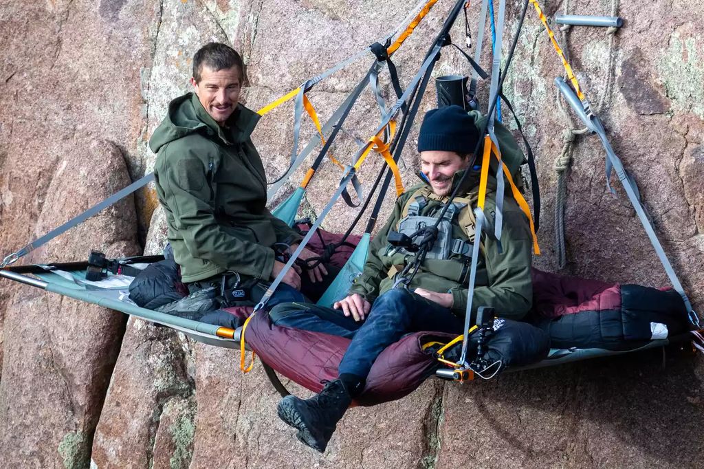Bradley Cooper and Bear Grylls sit on a swing bed on the mountain side in Wyoming
