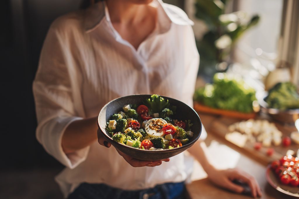 Healthy dinner or lunch. Woman in t-shirt and jeans standing and holding vegan superbowl or Buddha bowl with hummus, vegetable, salad, beans, couscous and avocado and smoothie in hands, square crop