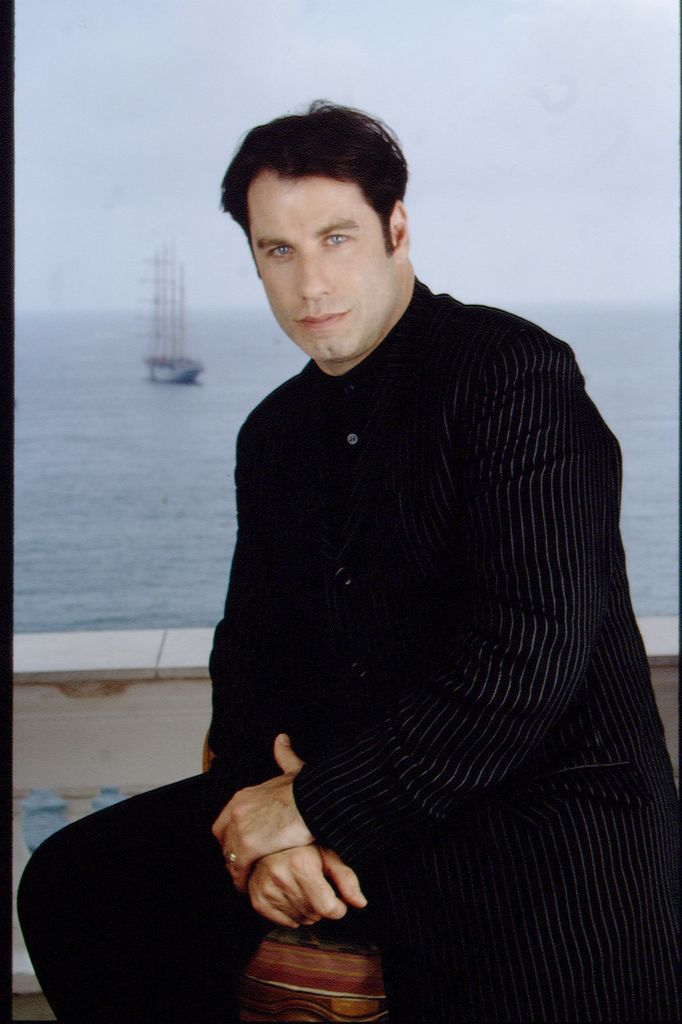 John Travolta est assis sur un tabouret, les mains jointes, vêtu de noir et sur fond de mer.