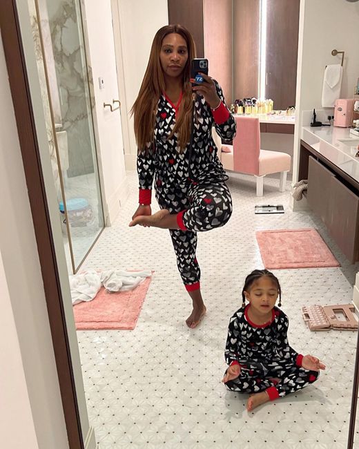 a photo of serena and her little girl in a large bathroom as they each perform a yoga pose one seated one standing on one leg and they both wear black pyjamas that are covered in white hearts with red stripes