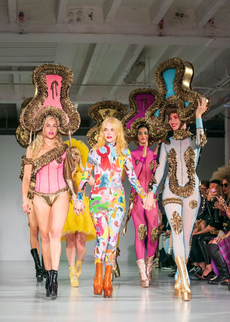 Pam Hogg a défilé lors du défilé SS20 de la Fashion Week de Londres dans une combinaison catsuit fluo brillante. Elle tient la main d’un mannequin portant un ensemble néon et un casque géant en guirlandes.