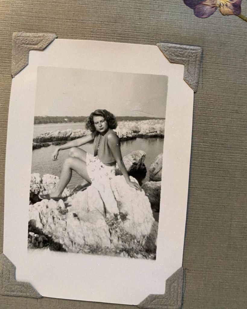 A black-and-white photo shows Lily James’ French grandmother as a young woman, sitting on rocks by a tranquil shoreline in a floral sundress. The image channels 1950s Riviera glamour and family legacy in its purest form.