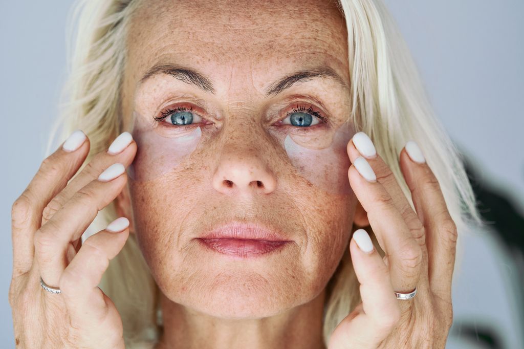 Older woman applying on her face hydrogel under-eye recovery patches. Do morning routine, to reduce puffiness. Skincare treatments usage, beauty routine concept