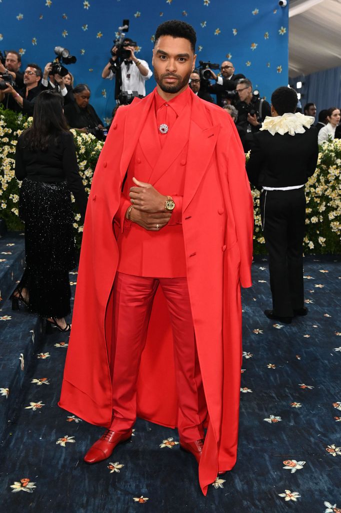 Rege Jean Page wears a bright red suit and long coat with matching red shoes