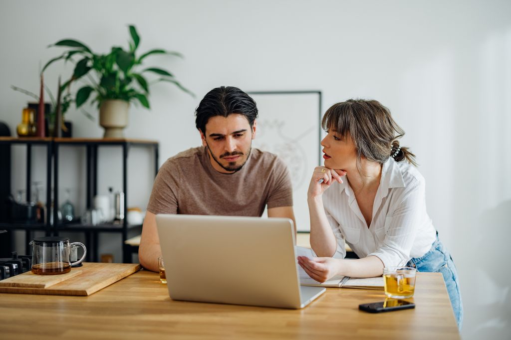 Boyfriend and girlfriend talking and doing home finances together online on a laptop computer in the kitchen.