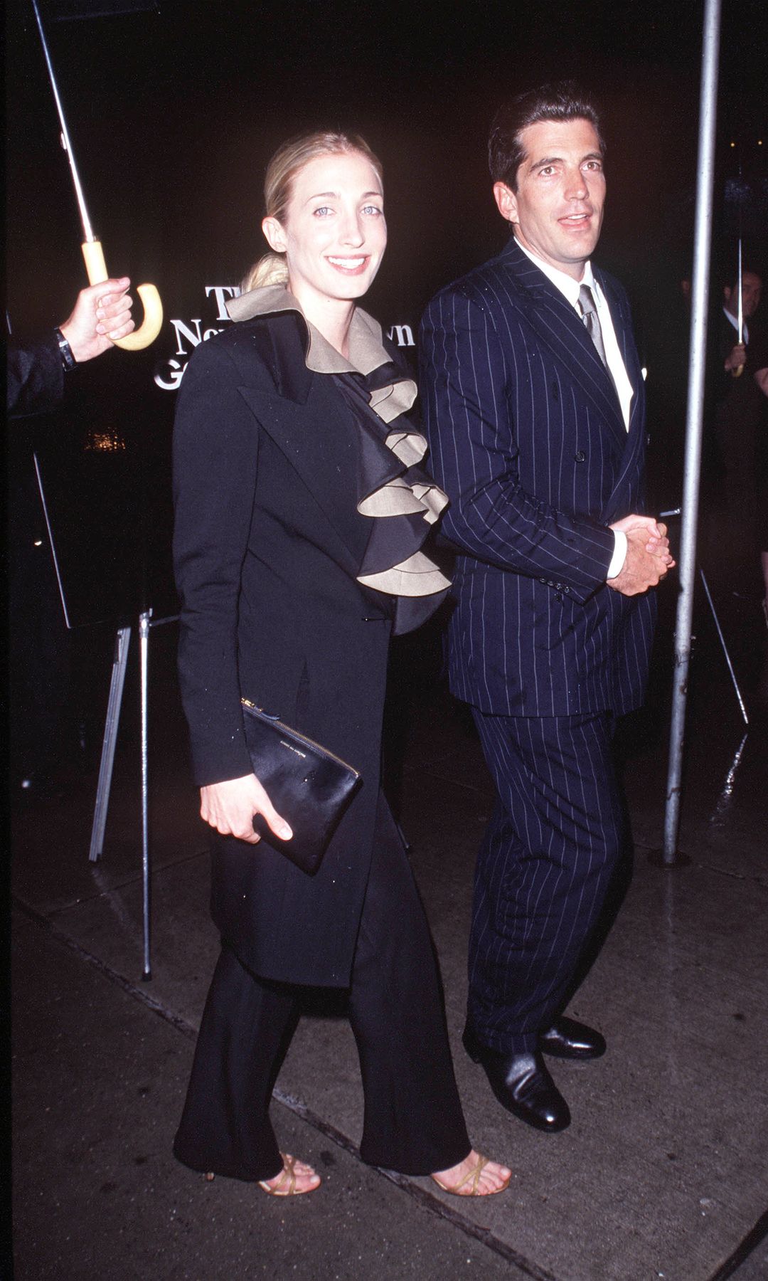 Carolin Bessette-Kennedy and John F. Kennedy Jr. walk down the street in New York, Carolyn wears a black and tan ruffled suit and John wears a pinstripe suit