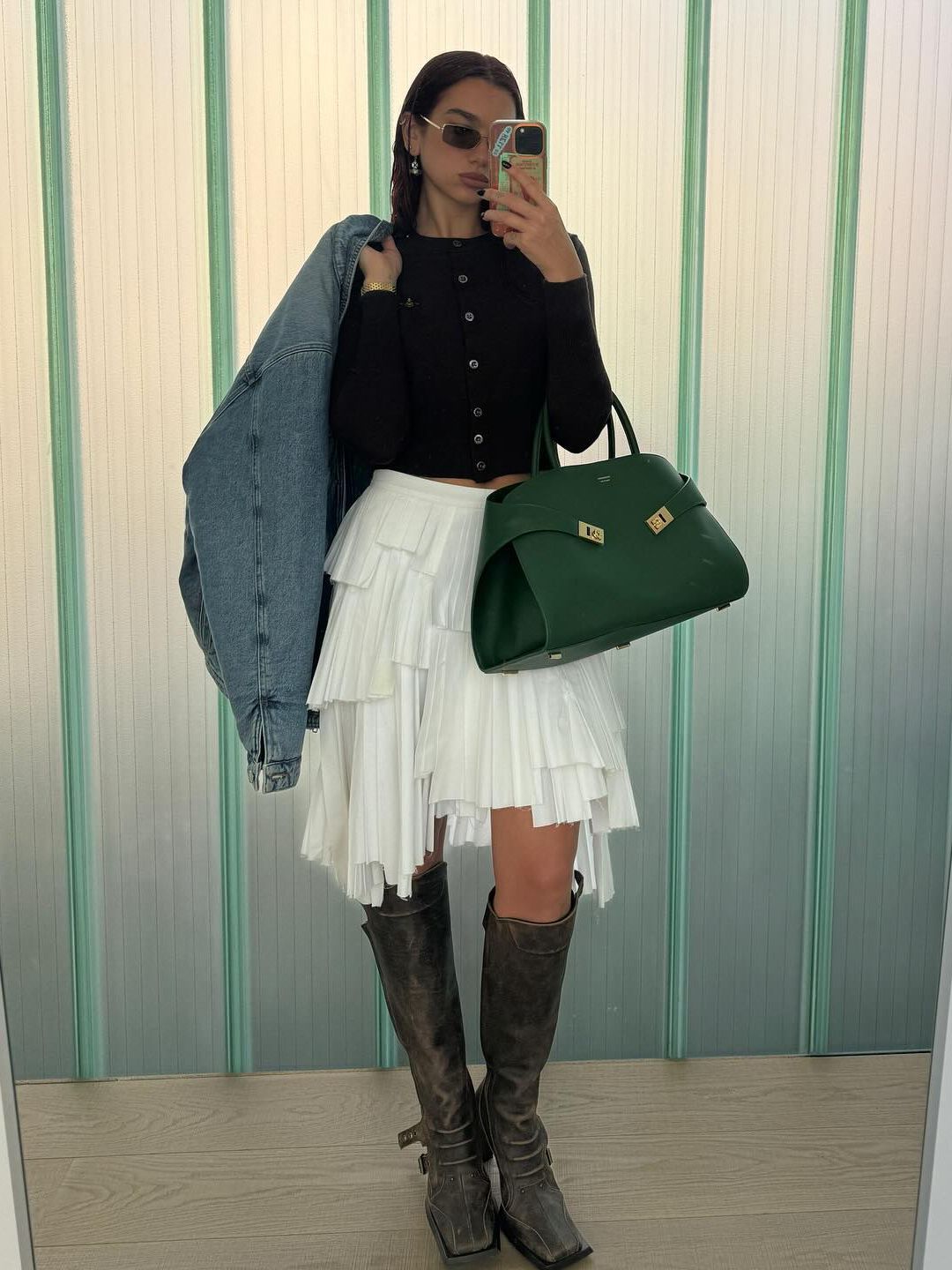 Dua Lipa poses for a mirror selfie in a white skirt, knee-high boots and knitted cardigan before heading off to work