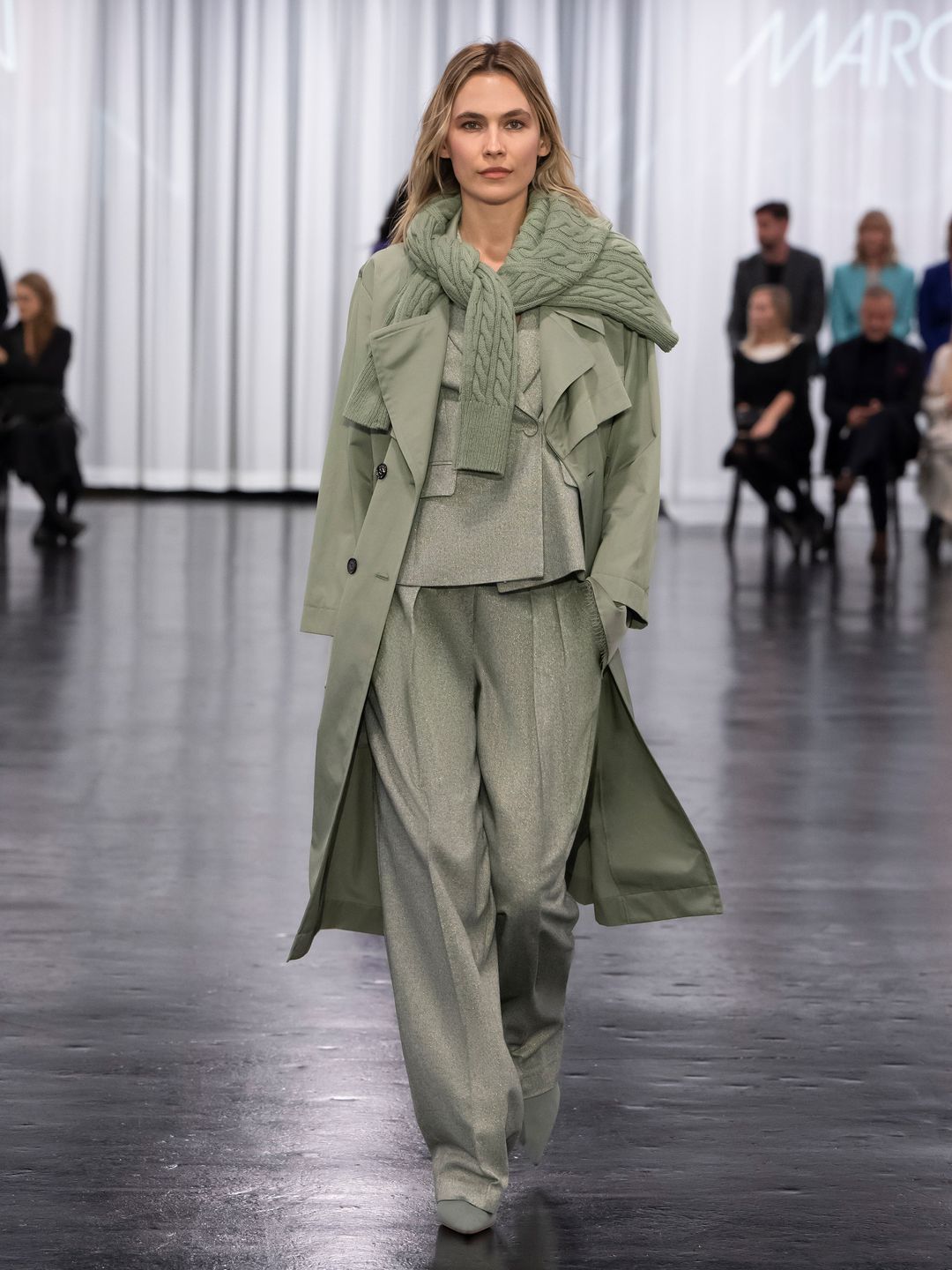 A Model at Marc Cain wears a sage green matching suit, trench coat, boots and sweater 