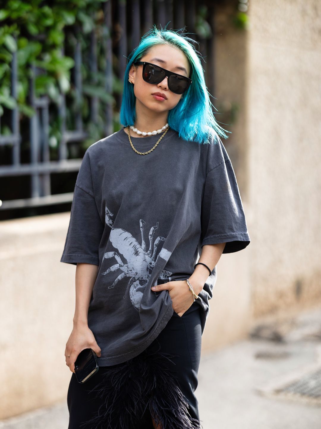 Margaret Zhang is seen wearing blue hair, black sunglasses, a pearl necklace, a gold necklace, yellow and black leather heels, an oversized grey t-shirt and a long black skirt embellished with feathers outside the Max Mara show during the Milan Fashion Week 2024