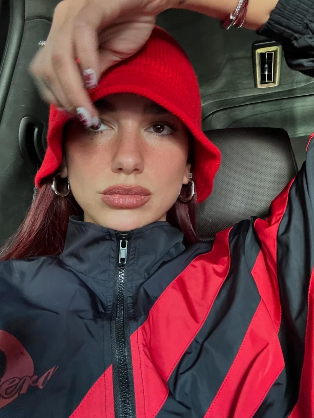 Dua Lipa poses in a car wearing a black and red jacket, a red hat and silver hoop earrings