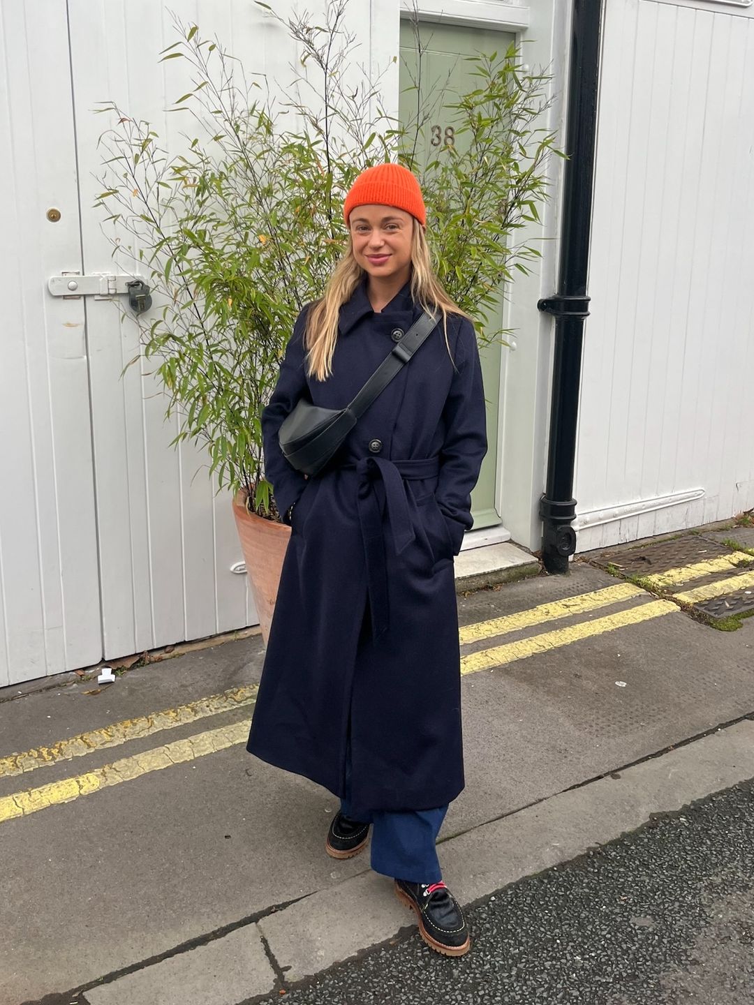 Amelia Windsor poses on the street in a navy trench coat, jeans, loafers and an orange beanie