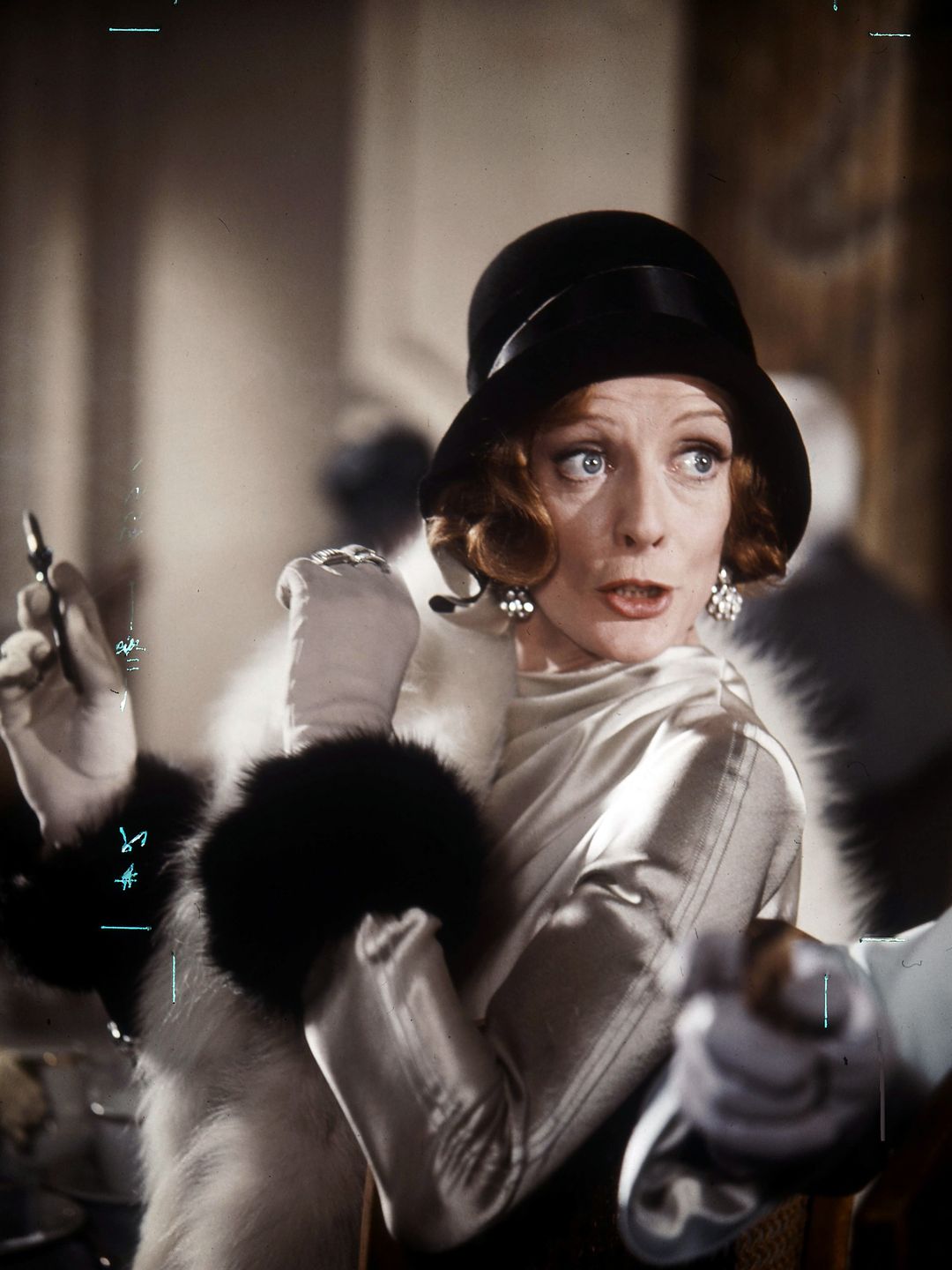 In this shot, Maggie Smith is dressed in an elegant satin gown, with long gloves and a luxurious fur stole wrapped around her shoulders. She wears a black hat, and her short hair is curled beneath it. Her expressive face is caught mid-conversation, looking slightly surprised, with wide eyes. The scene exudes 1920s-style glamour, evoking a sense of refined, classic beauty.