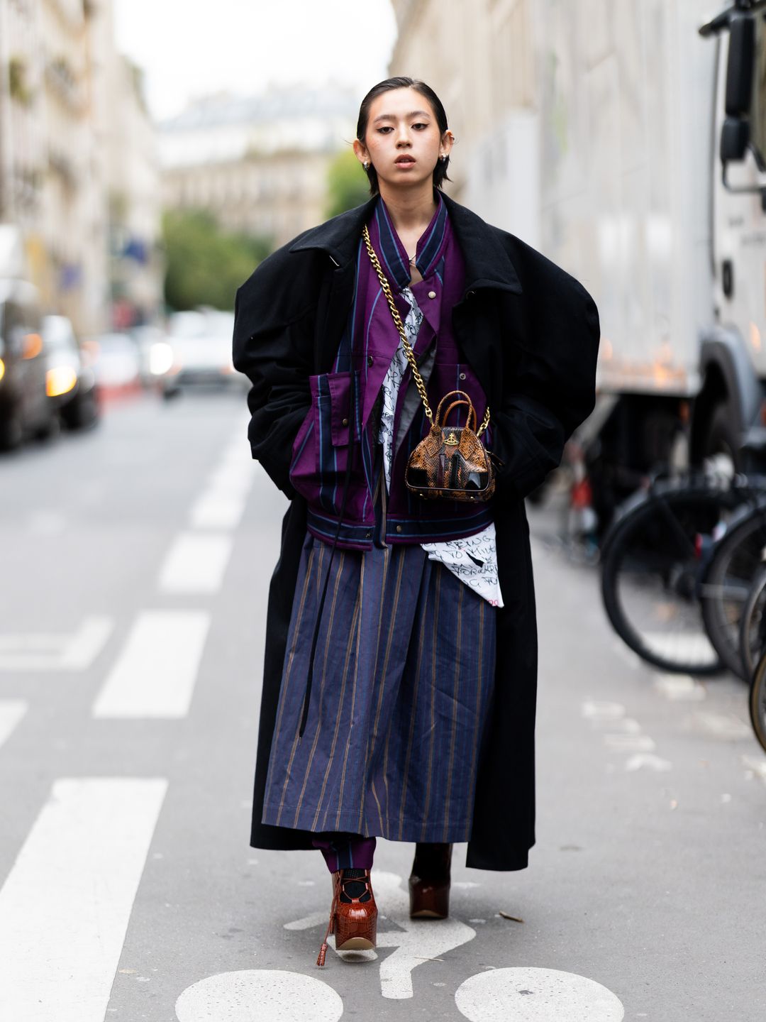 Tan Suan wears a navy and pink pinstripe skirt in Paris, paired with silver earrings, an oversized black coat, a purple and blue bomber jacket and leather platform heels outside Vivienne Westwood.