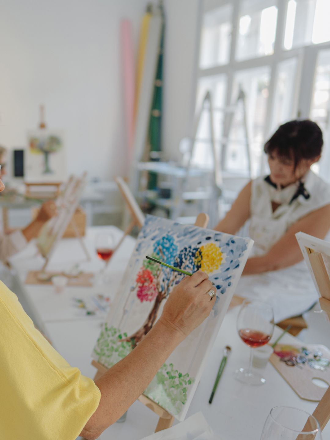 Photo of a woman taking painting classes with her friends in a local art studio; connecting with her friends over a glass of wine and learning how to paint together.
