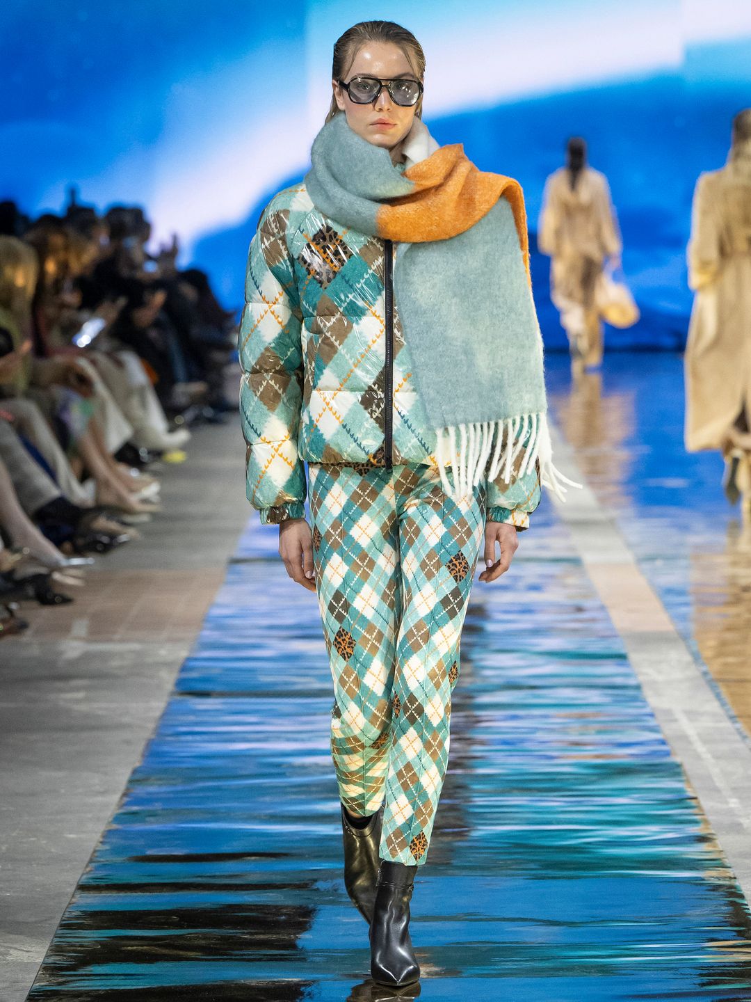 A model walks the runway for Marc Cain wearing a matching argyle print padded jacket and trousers in white, green, brown and leopard print. She wears a blue and orange scarf draped around her neck, black boots and black sunglasses. 