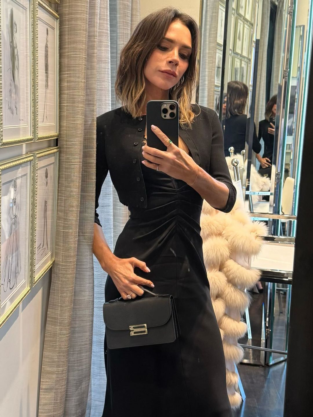 Victoria Beckham poses for a mirror selfie in a black dress, cropped cardigan and black clutch bag