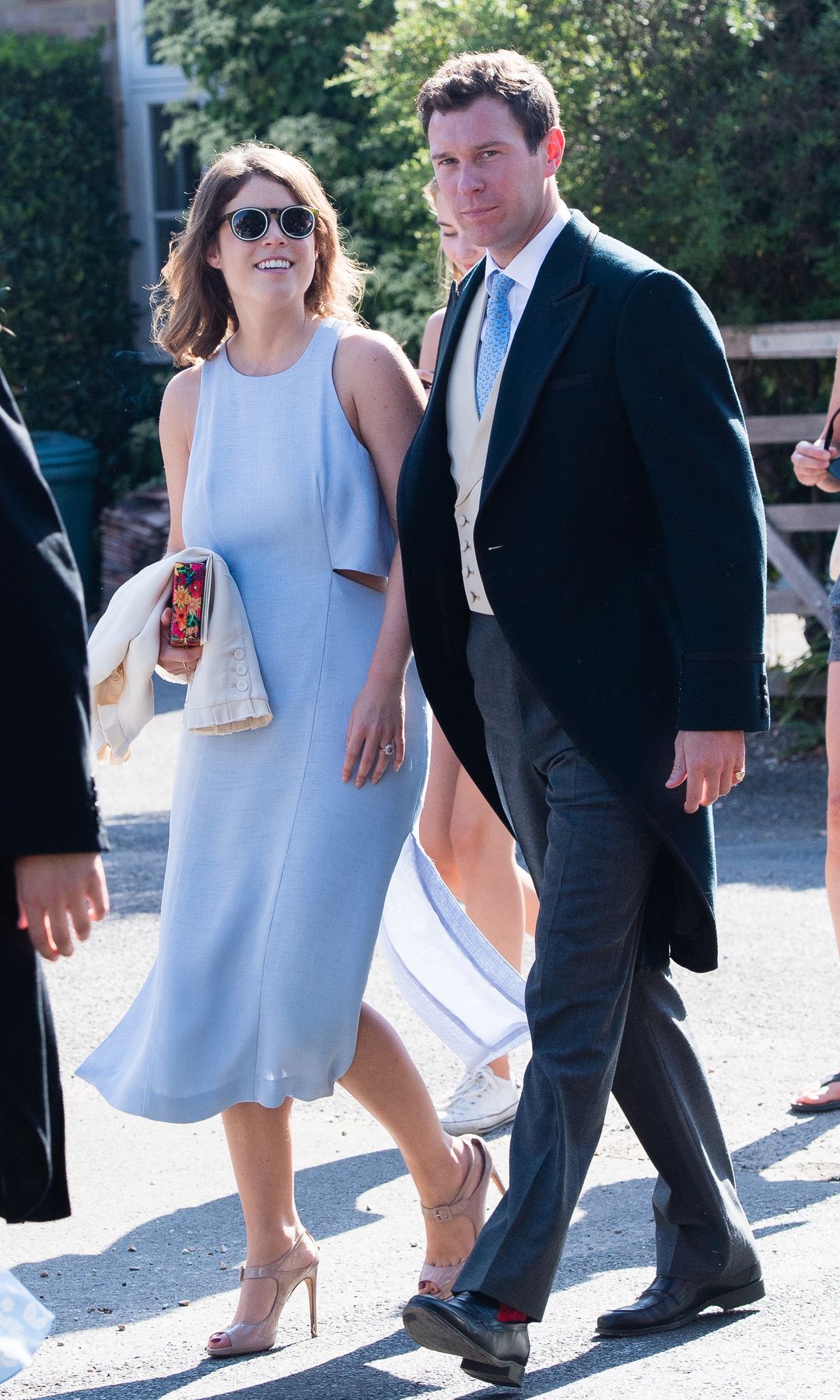 Princess Eugenie and Jack Brooksbank walk through the church yard, Eugenie wearing a pale blue high neck midi dress with pale heels and black sunglasses
